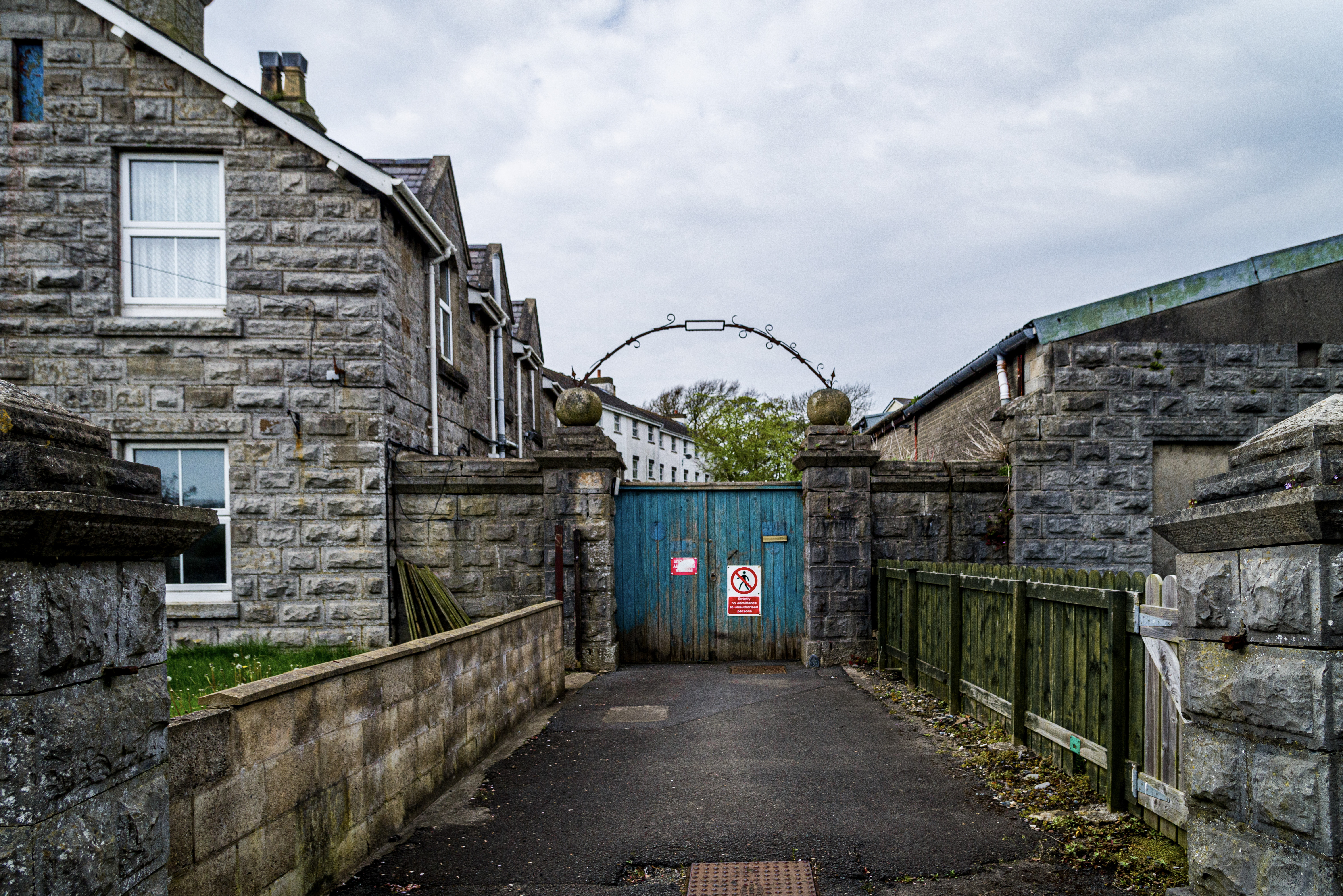 A gritty scene featuring a large blue painted gate at the end of a driveway barring access to an old stone-brick property.