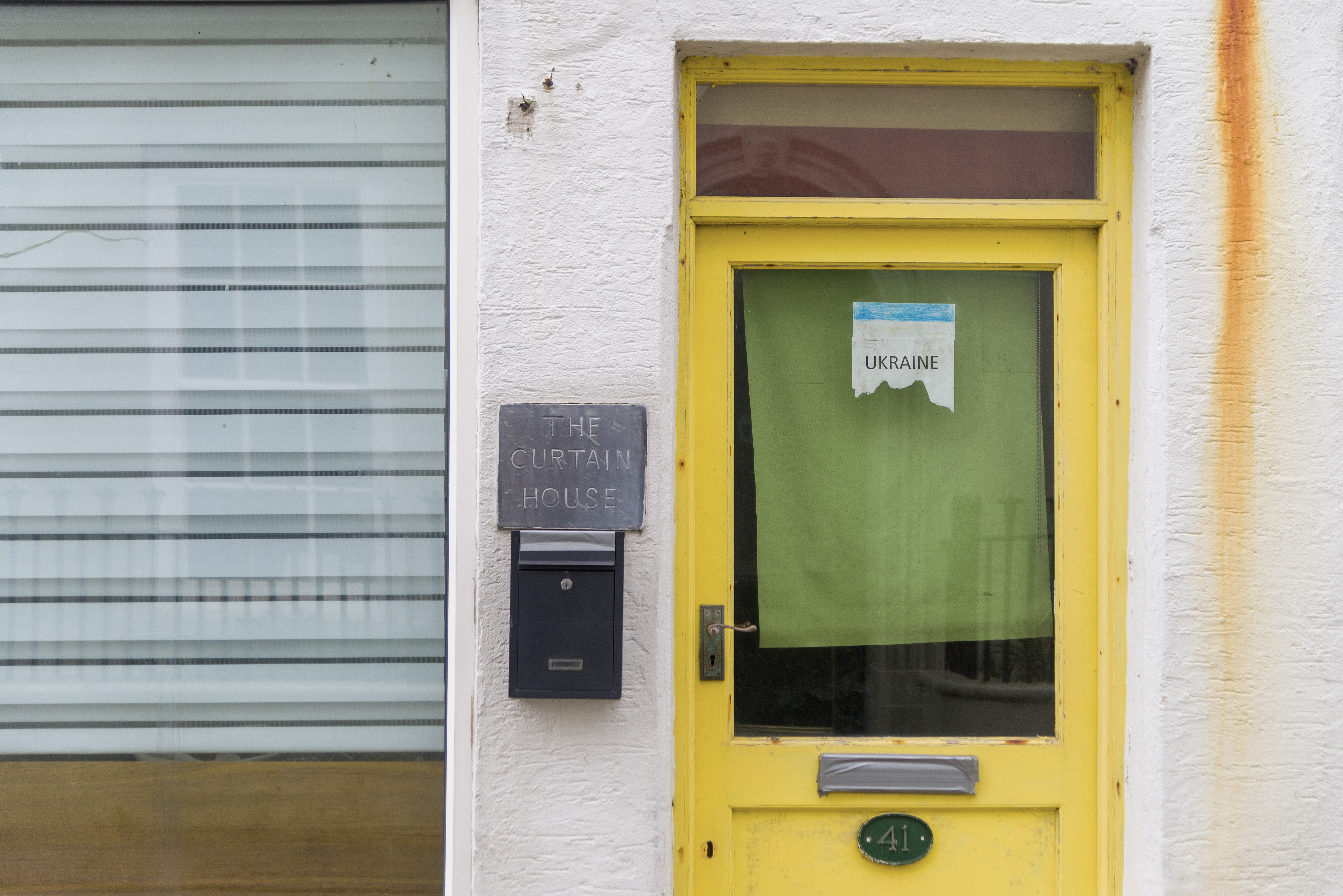 A yellow door with a glass window, on the window is a torn paper bearing the word “Ukraine” and behind that is a green blind. To the left of the door is a plaque containing the text “The Curtain House”, to the left of the plaque is another window and in that window is a another blind.