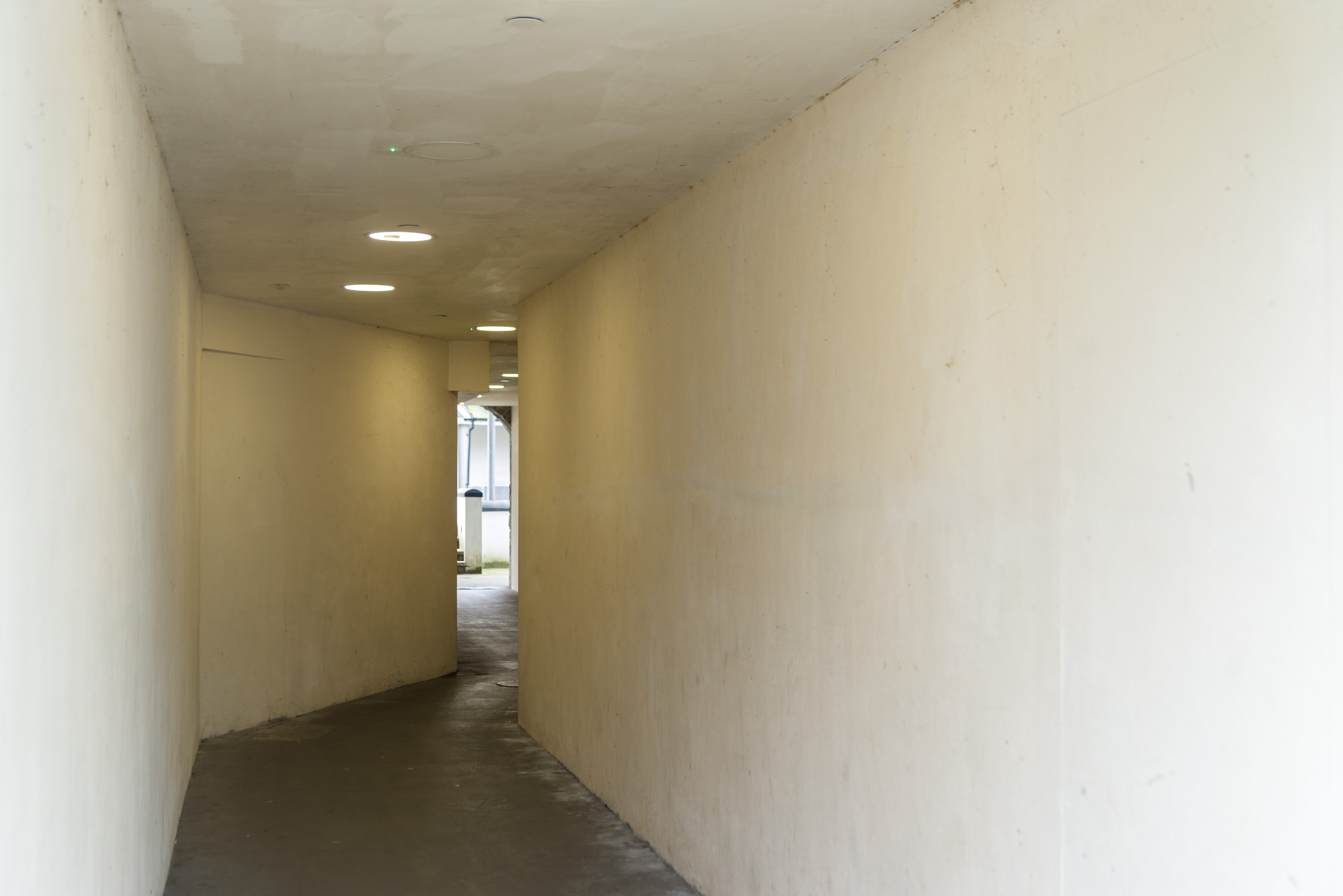 A passageway snakes through a building revealing a small glimpse of the courtyard beyond. It is drab and eerie in it’s design.