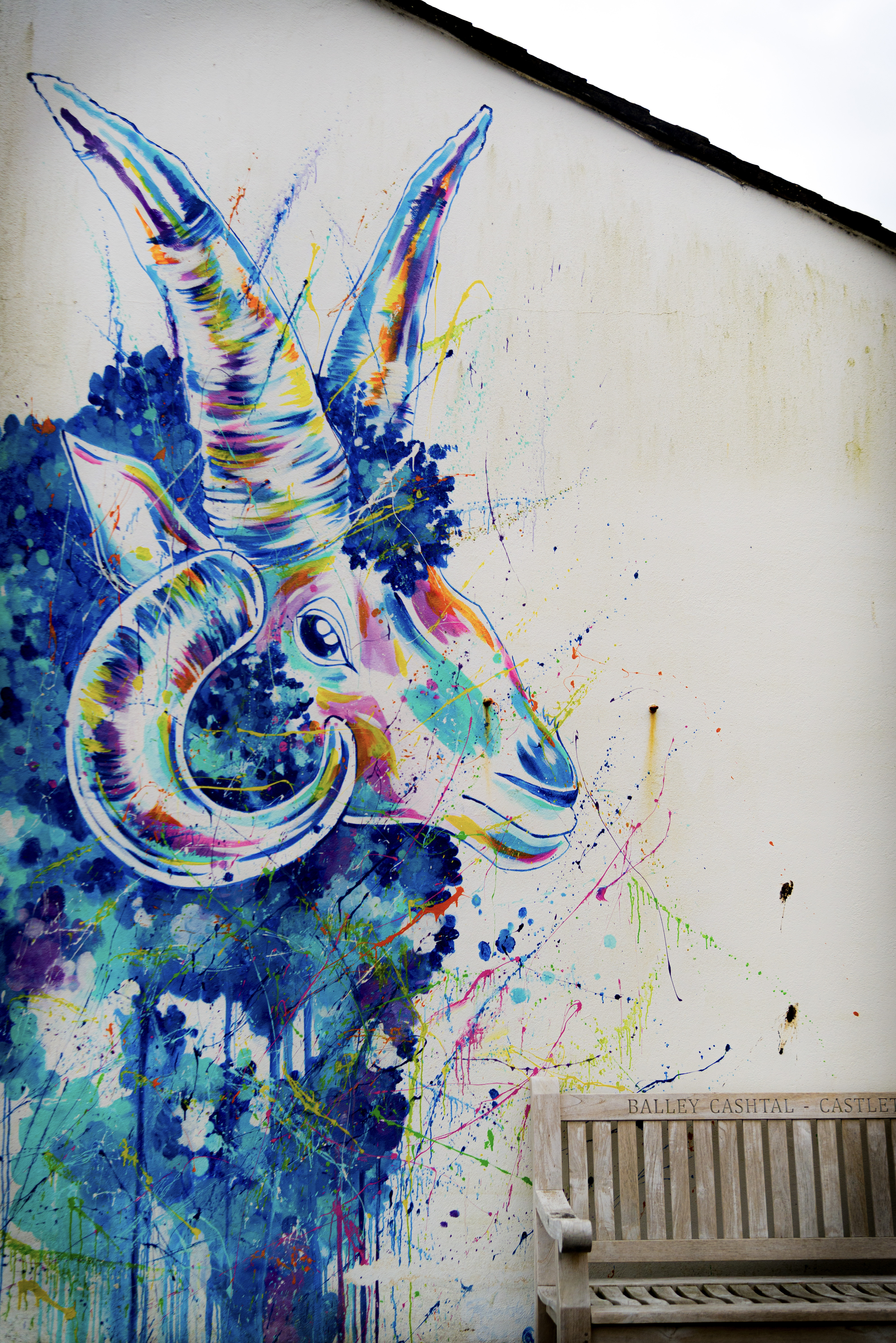 A mural of a loaghtan sheep painted on the side of a building, to the bottom right sits a bench.