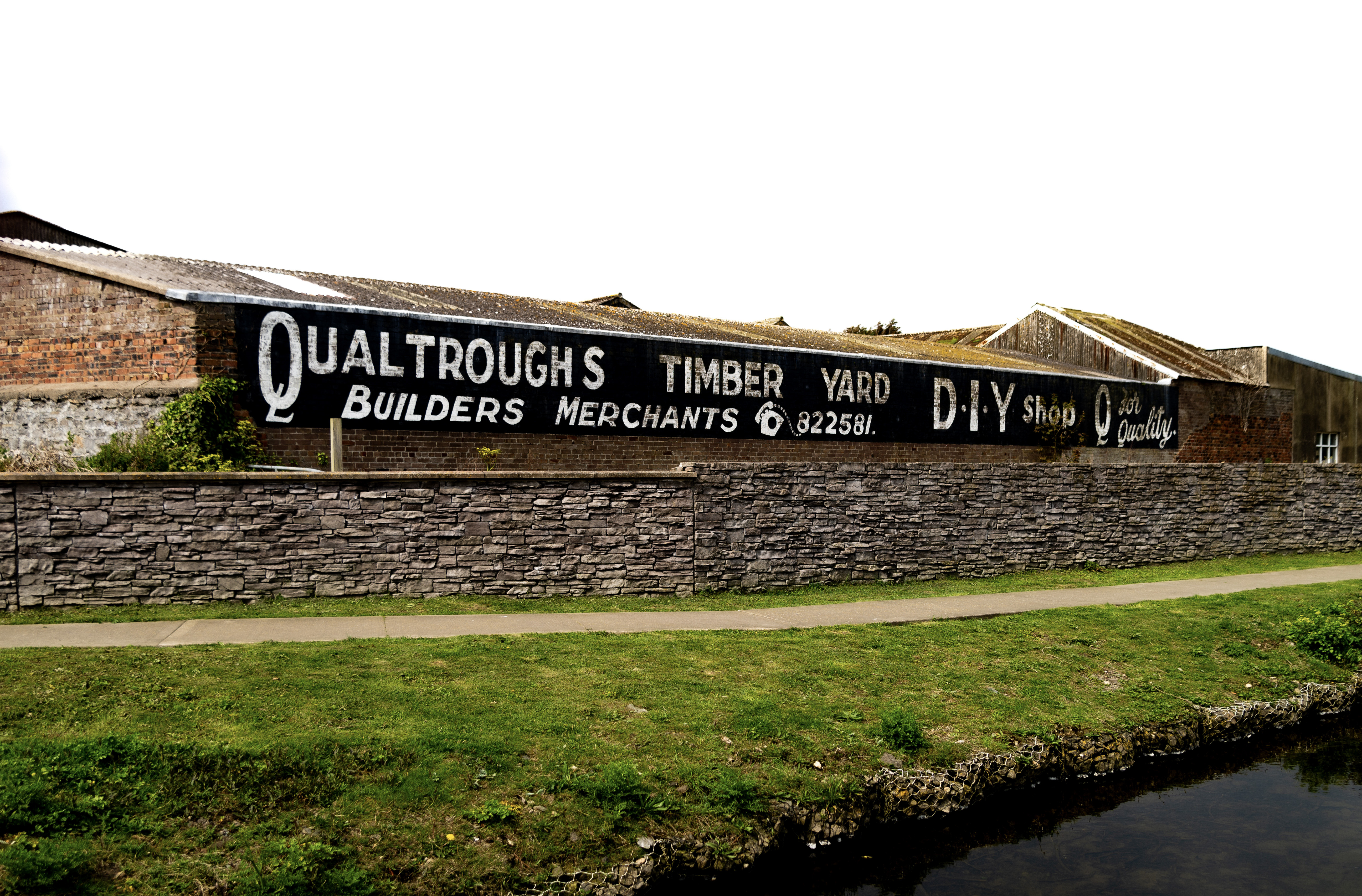 A large sign reads “Qualtroughs Timber Yard D.I.Y. Shop Q for Quality”, it is advertising a builders merchants. A river path makes up the foreground of the scene, and an old stone wall stands between the builds merchant and the path.