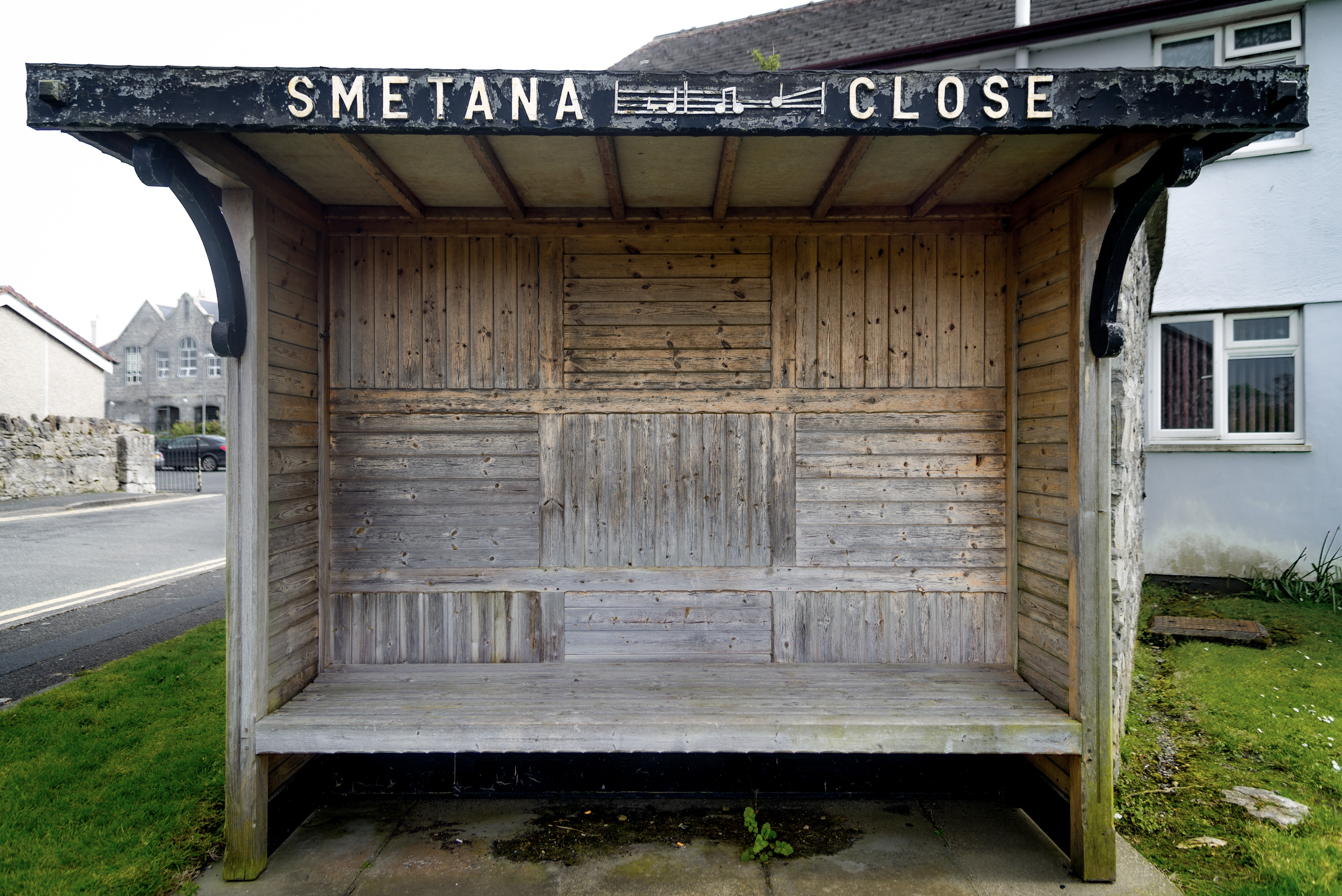 An old wooden bus stop with the words “Smetana Close” on it’s roof. The wooden slates alternate in a grid pattern, and between the words is a musical score.