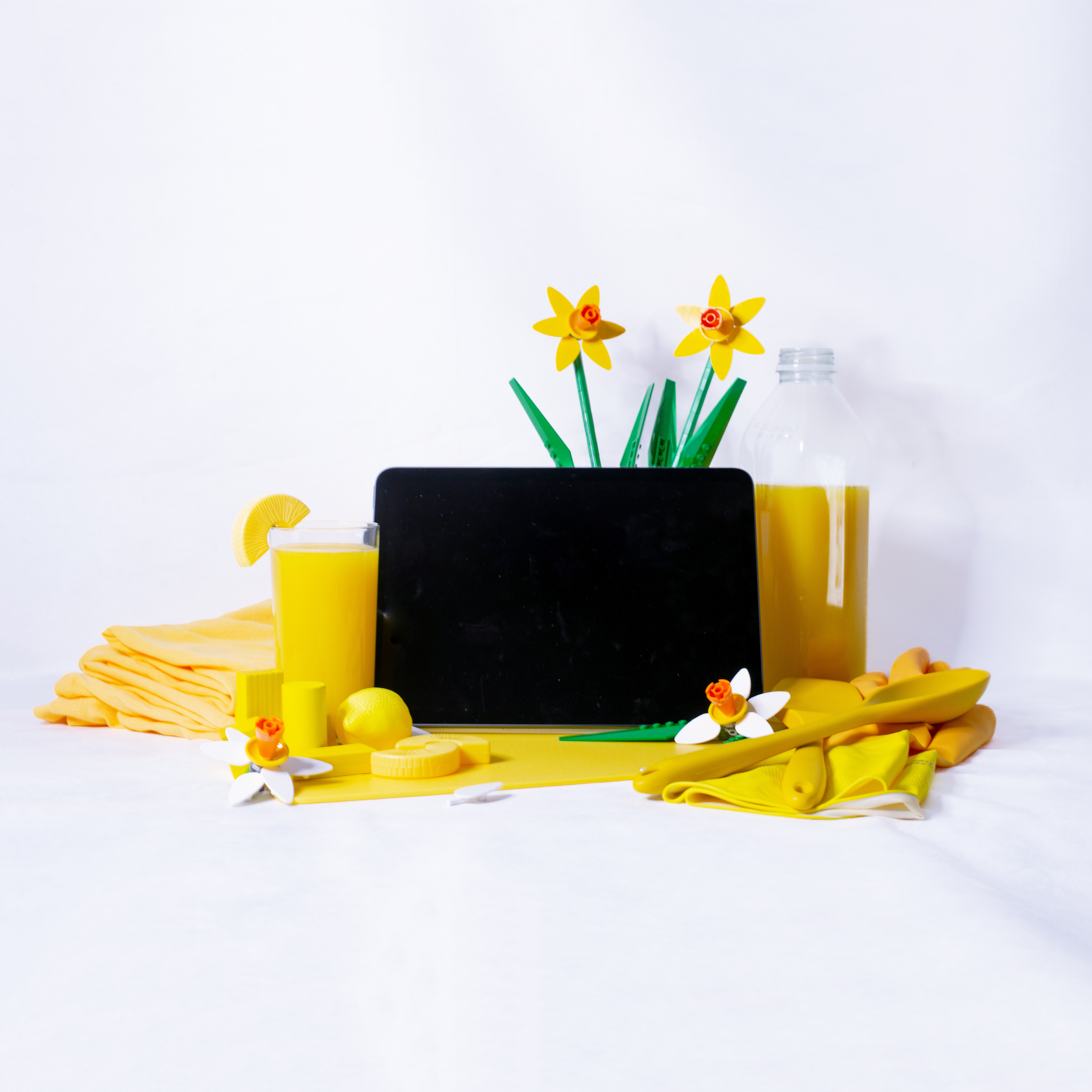 A tablet, it’s screen black, is nestled between some marigold cleaning gloves and some orange juice. Beneath the tablet is a yellow cutting board, and around it are yellow plastic food and yellow playing blocks. Behind the tablet are some lego daffodils.