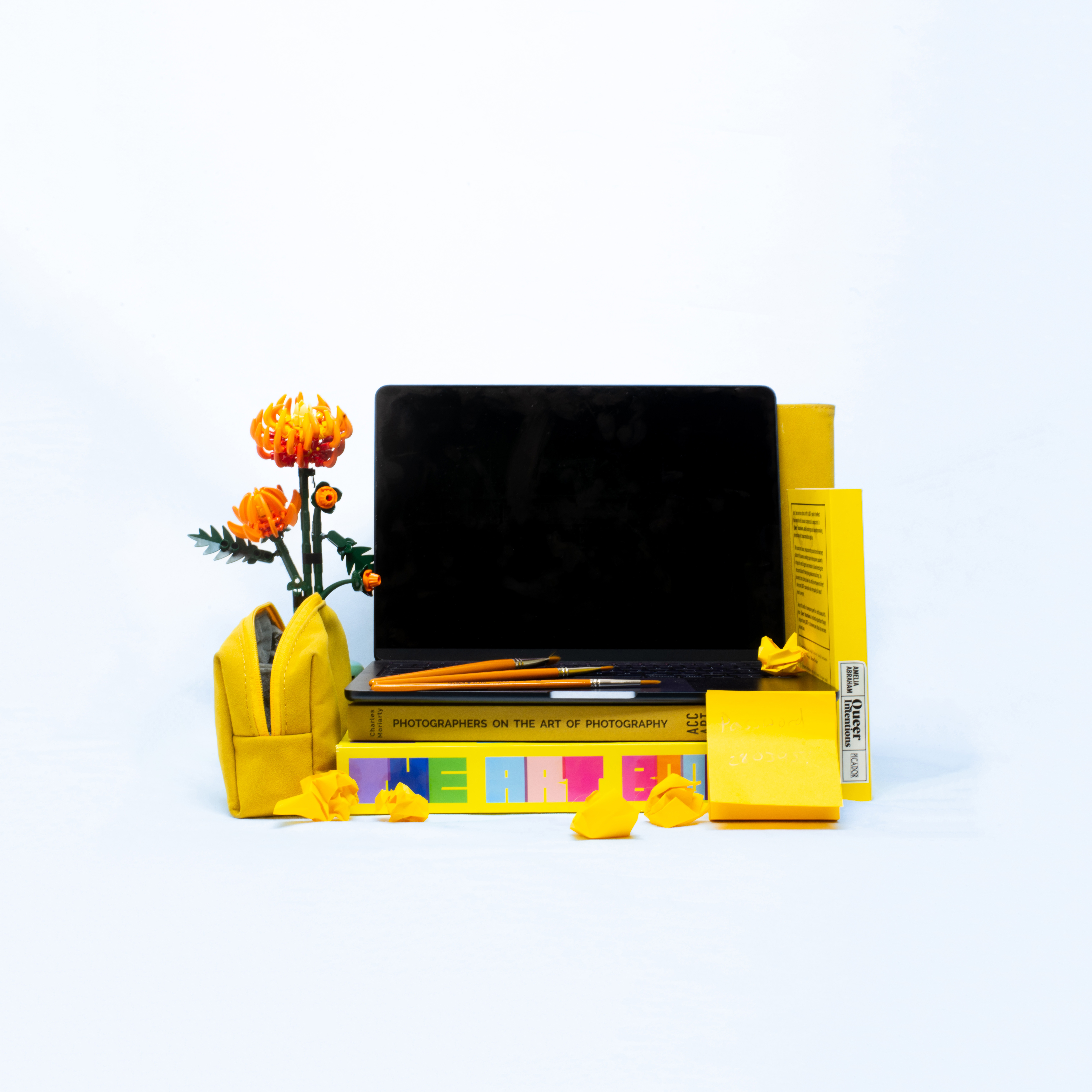 A laptop, it’s screen black, is standing open on a pile of yellow art books surrounded by yellow creative tools. A lego crystanthemum stands to it’s left.